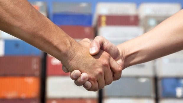  The image depicts a firm handshake between to parties with blur background of shipping containers to showcase Cross-sector collaboration